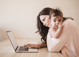 A mom holding her infant looks at and types one-handedly on her laptop.