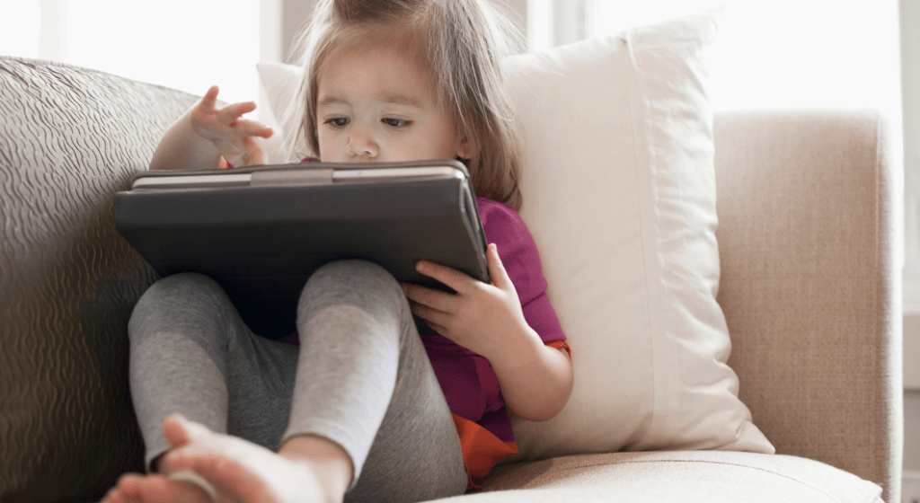 Little girl with a tablet sits on the couch alone.