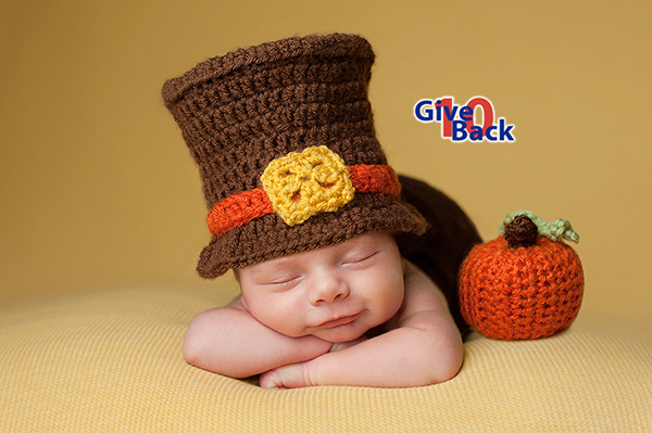 Smiling four week old newborn baby boy wearing a crocheted Pilgrim hat. He is sleeping on a gold blanket next to a crocheted pumpkin.