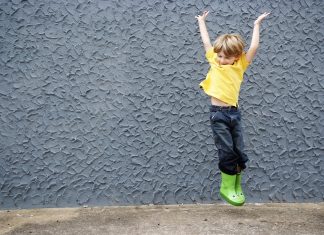 A boy jumps in a pair of green froggy rain boots.
