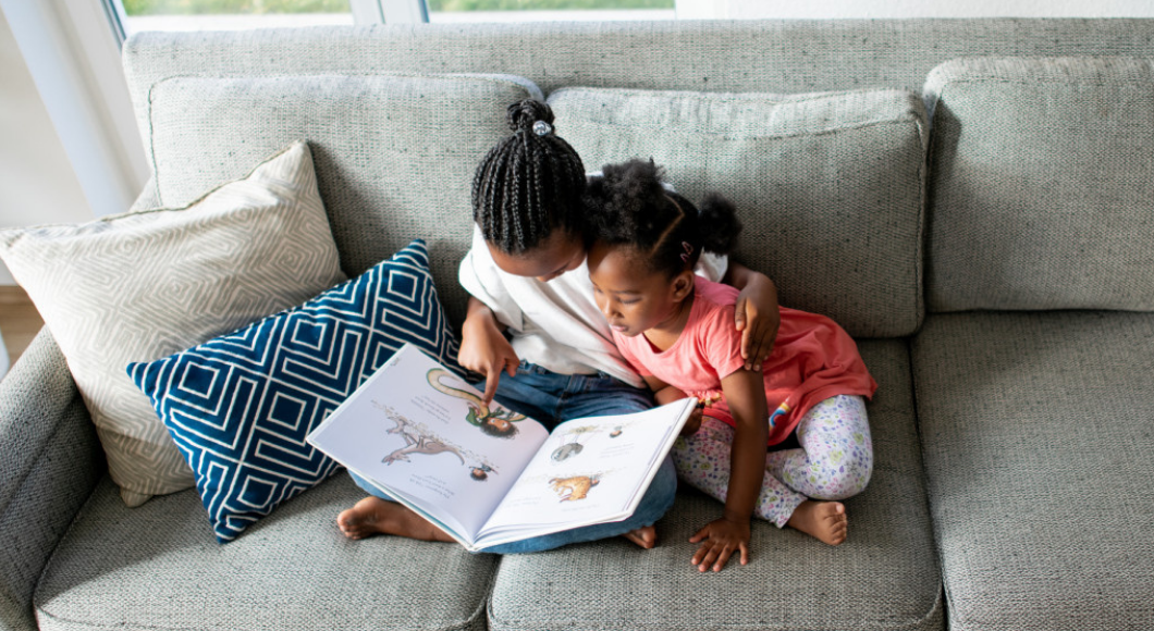 Two girls read a book on a couch.