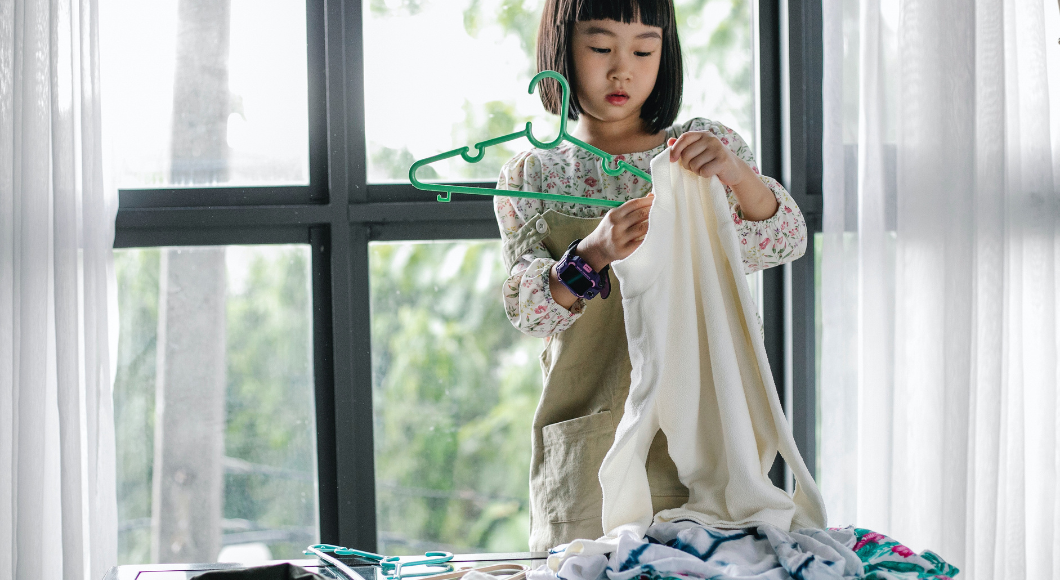 A little girl puts a shirt on a green hanger, helping with the laundry and chores.