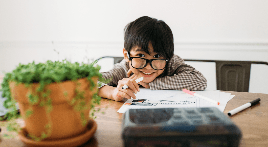 smiling child working on homework with a plant and pencil box in the foreground