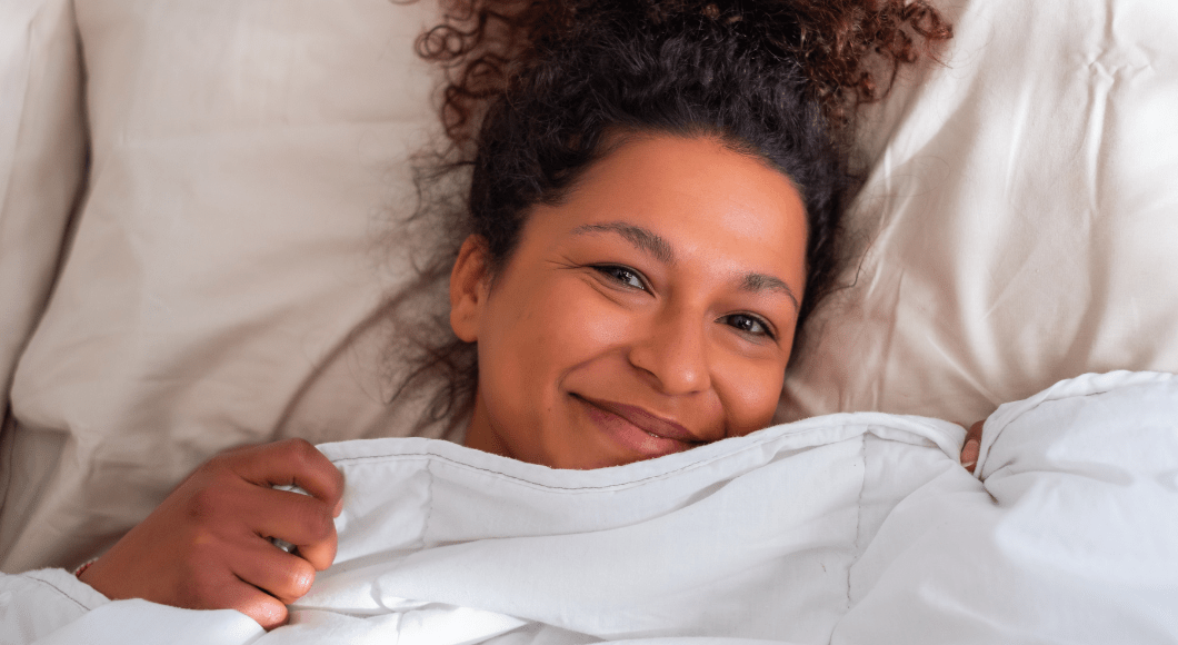 tired woman smiling in bed with covers pulled up to her chin