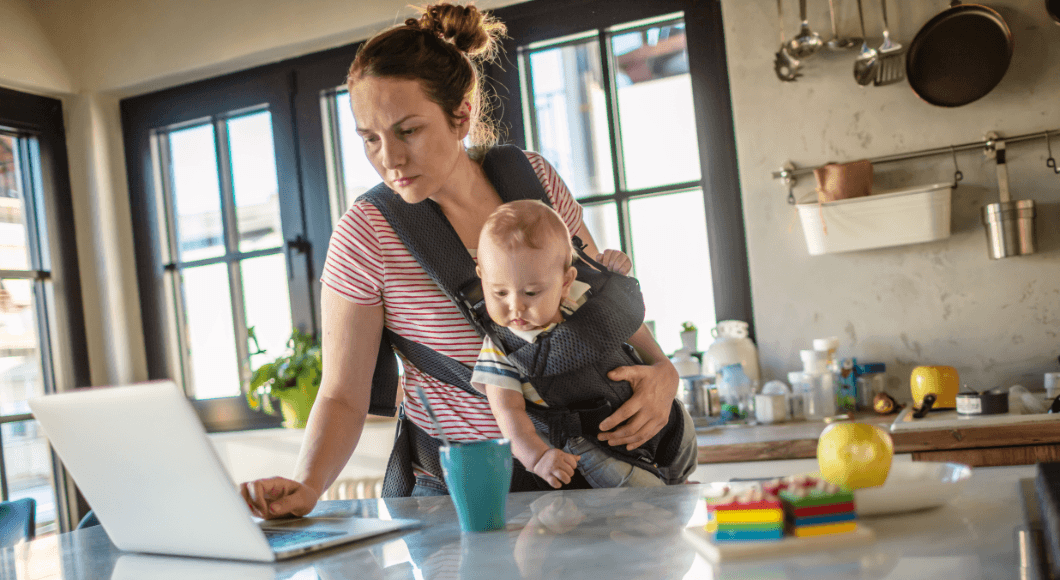 mother standing in the kitchen working on a laptop while holding an infant in a baby carrier