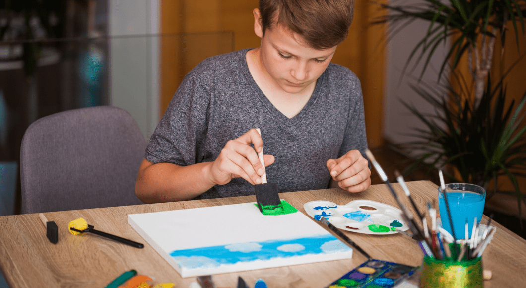 A tween boy sits at a table at home and paints on a canvas.