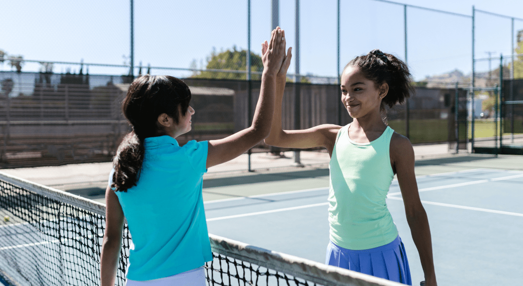 Two young girls high-five each other over a tennis net.