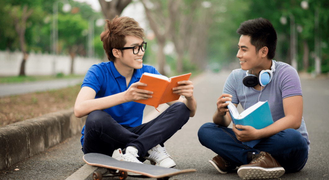 Two teens read books. One sits on a skateboard.