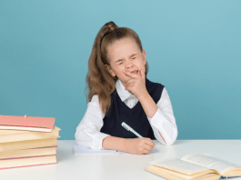 Young girl in a school uniform with books at a desk closes her eyes and squishes her face as if she is upset