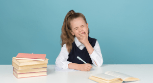 Young girl in a school uniform with books at a desk closes her eyes and squishes her face as if she is upset