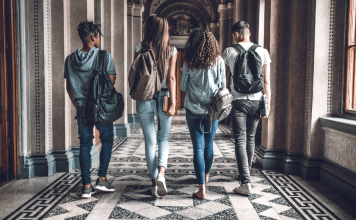 Two boys and two girls of college age walk down a tiled hall with their backs and backpacks