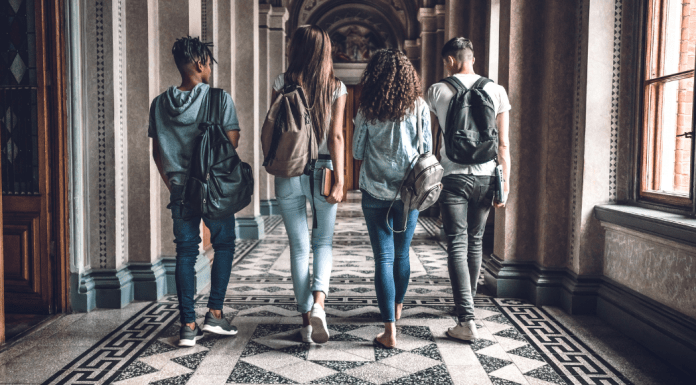 Two boys and two girls of college age walk down a tiled hall with their backs and backpacks
