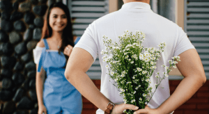 A man holds a bouquet of daisies behind his back to surprise his girlfriend standing in front of him.
