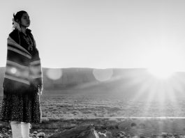 A Native American girl wrapped in a Navajo blanket with a sun beam behind her.