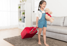 A girl wheeling a red suitcase and holding a teddy bear.