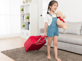 A girl wheeling a red suitcase and holding a teddy bear.