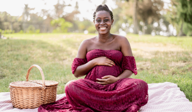 A pregnant woman wearing a maroon summer dress sits on a picnic blanket holding her belly.