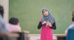 Young girl wearing a hijab stands at the front of her classroom showing a project to her classmates.