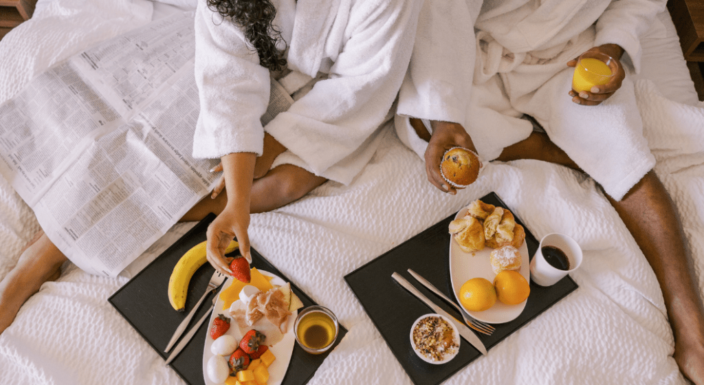 A couple wearing bathrobes eat room service in a hotel room.