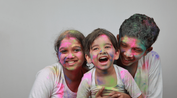 Three children smile, laugh, and embrace covered in colors from a Holi celebration.